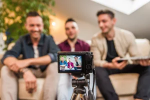 NSW regulators warn gambling companies over influencer marketing and online promotions. Three men sit on a couch while being filmed by a camera on a tripod, appearing to record a podcast or online video together.
