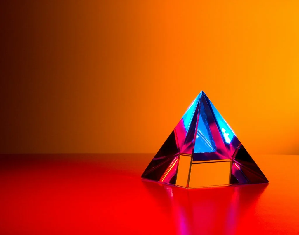 A geometric glass prism sits on a vibrant red surface against a saturated orange background. The prism refracts and reflects bright neon blue and magenta light, creating sharp, clean internal angles.